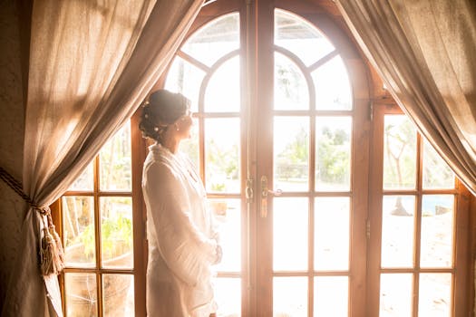 A woman standing by French doors, illuminated by soft morning light, creates a serene interior scene.