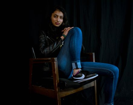 Portrait of a woman in casual fashion sitting on a chair in a dark studio setting.