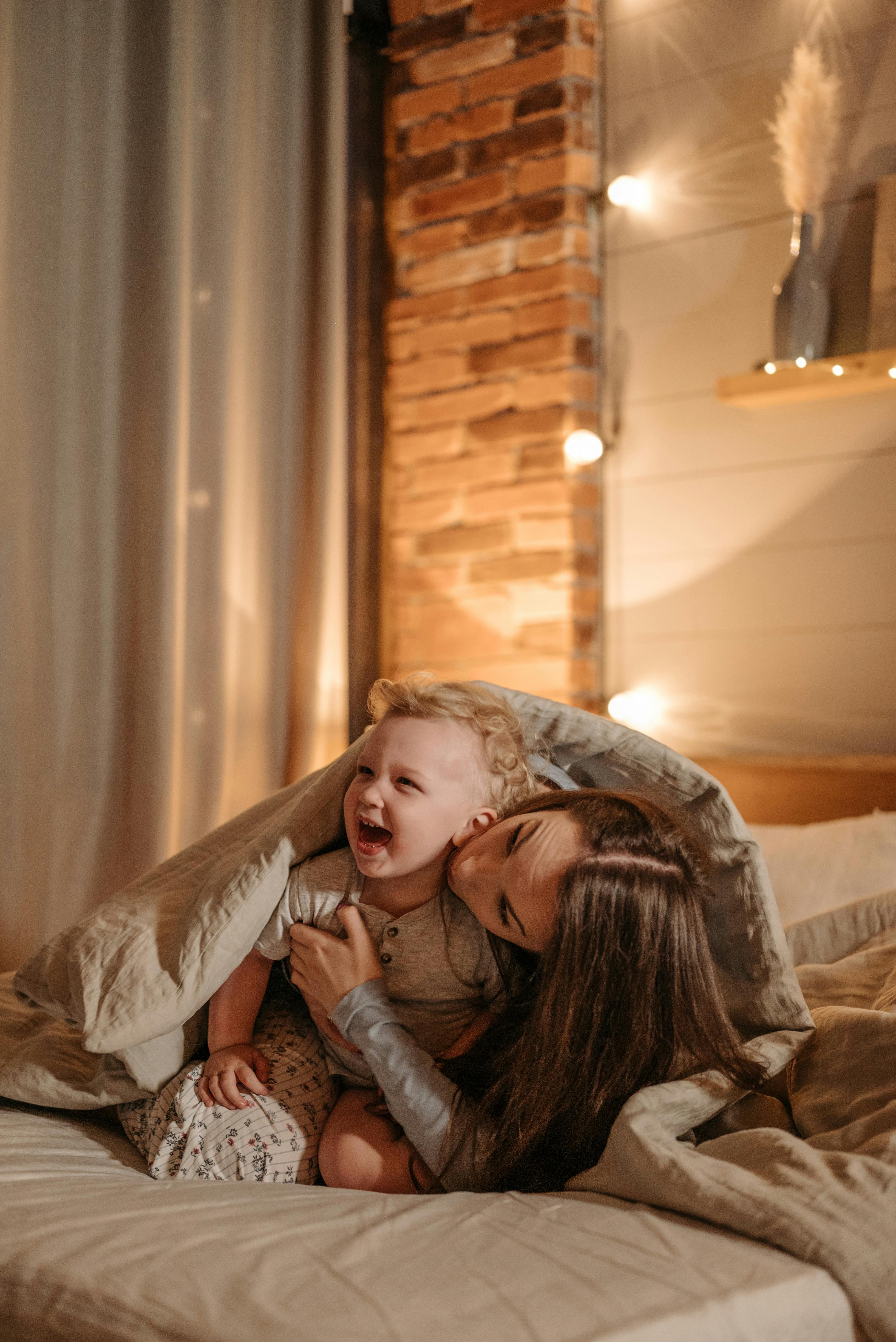 A mother and son share a joyful bonding moment on a cozy bed in a warmly lit bedroom.