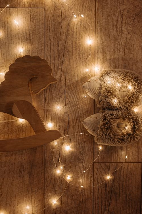 Free Cozy scene featuring a wooden rocking horse and fuzzy slippers decorated with warm fairy lights on a wooden floor. Stock Photo