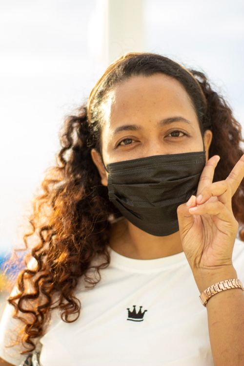 Free Portrait of a woman outdoors making a peace sign, wearing a black mask and white shirt. Stock Photo