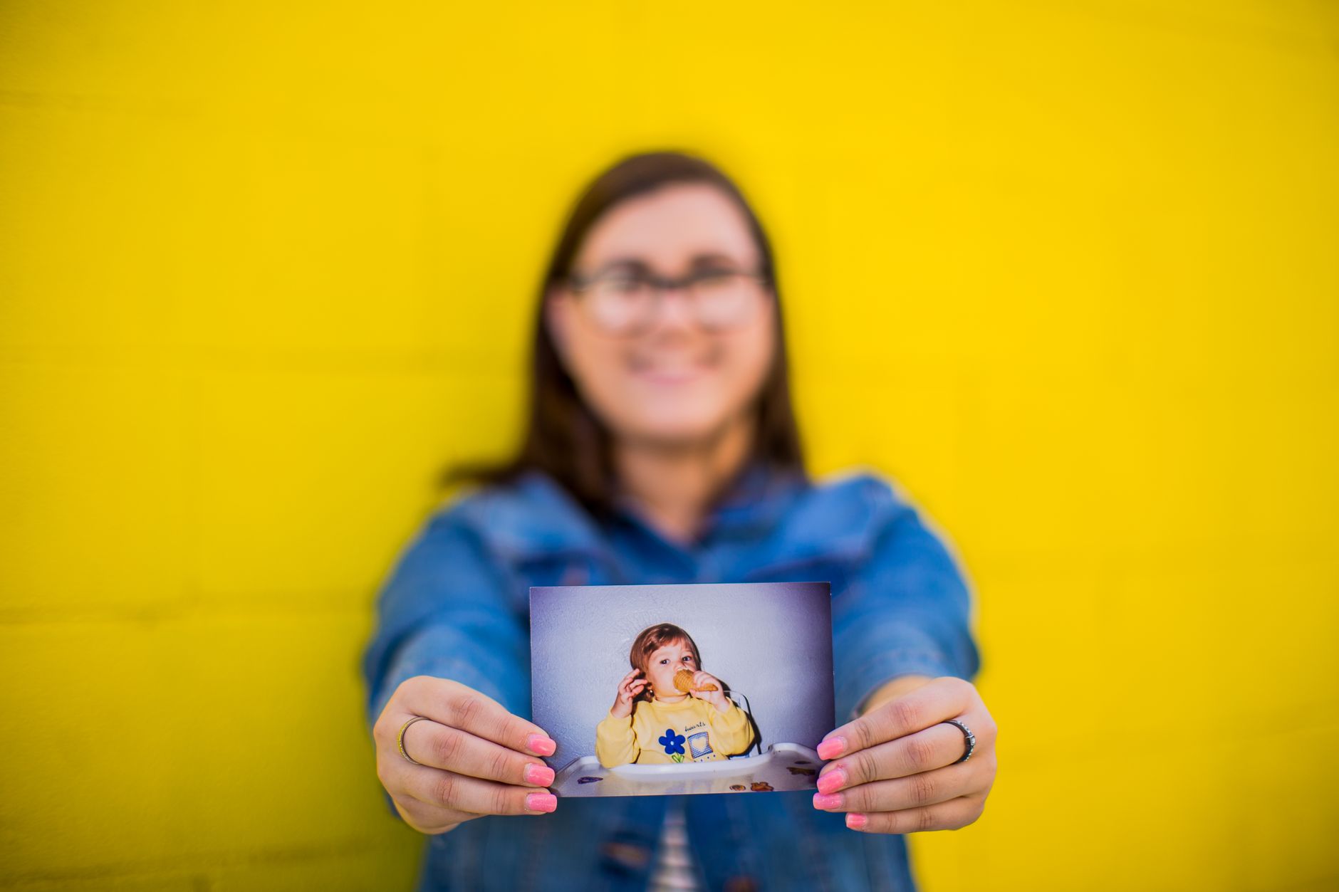 A smiling woman holding a nostalgic childhood photo against a vibrant yellow backdrop.
