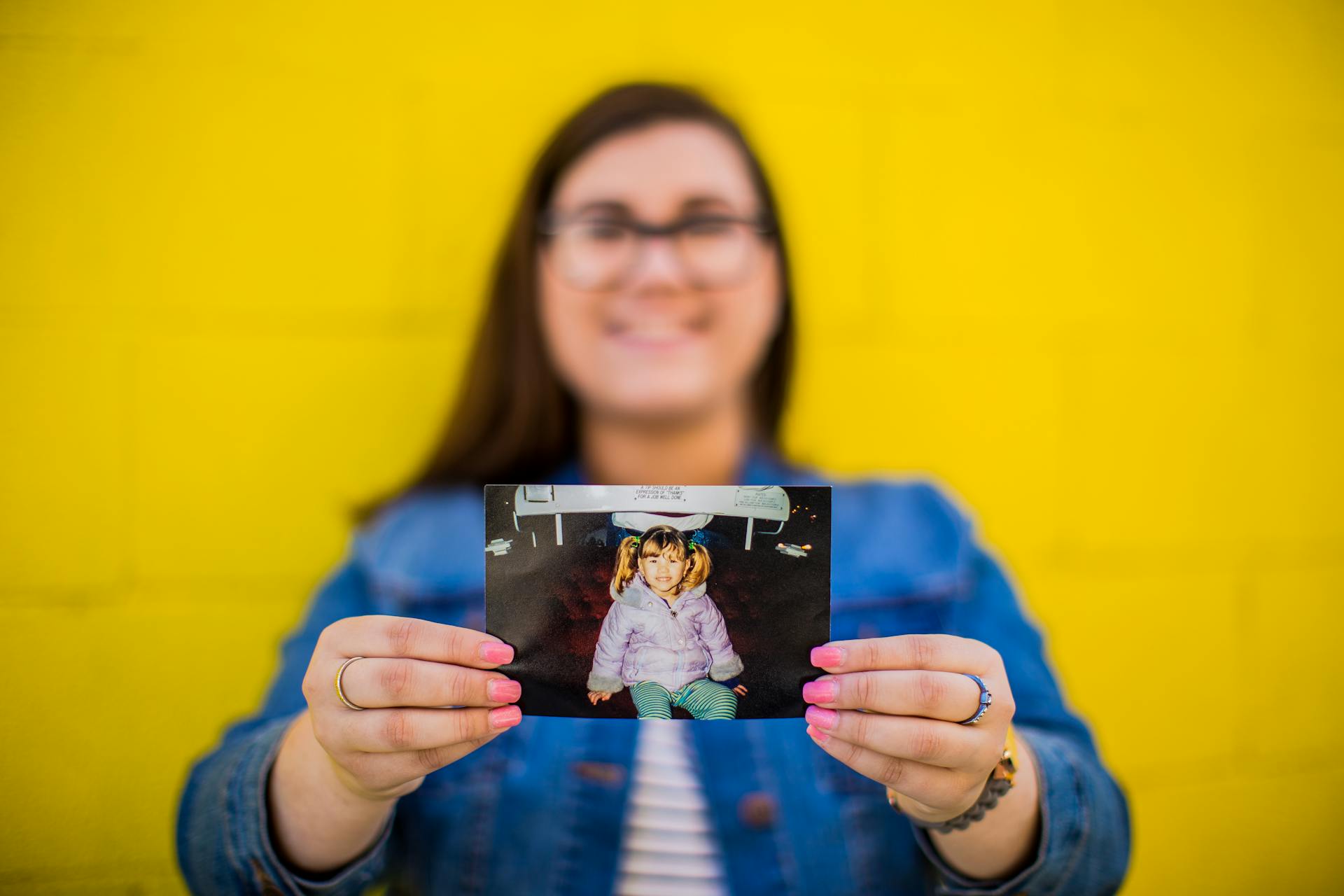 Blurred woman holding a childhood photo against a bright yellow wall background.