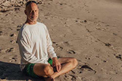 Free Man in White Long Sleeve Shirt and Green Shorts Sitting in Lotus Pose on Sand Stock Photo