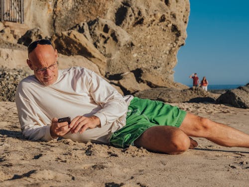Free Man in White Long Sleeve Shirt and Green Shorts Lying on the Side on the Beach and Using Mobile Phone Stock Photo