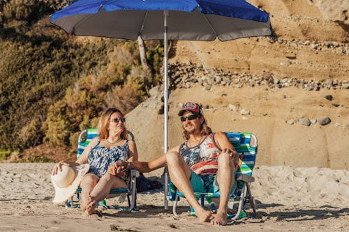 Free Woman in Blue Swimsuit and Man in Sunglasses Sitting Under Beach Umbrella and Holding Hands Stock Photo