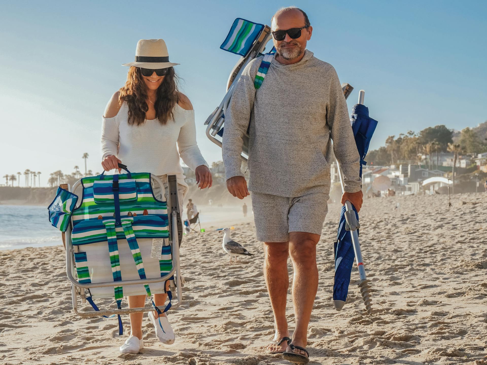 Man and Woman Carrying Foldable Chairs while Walking on the Sandy Shore