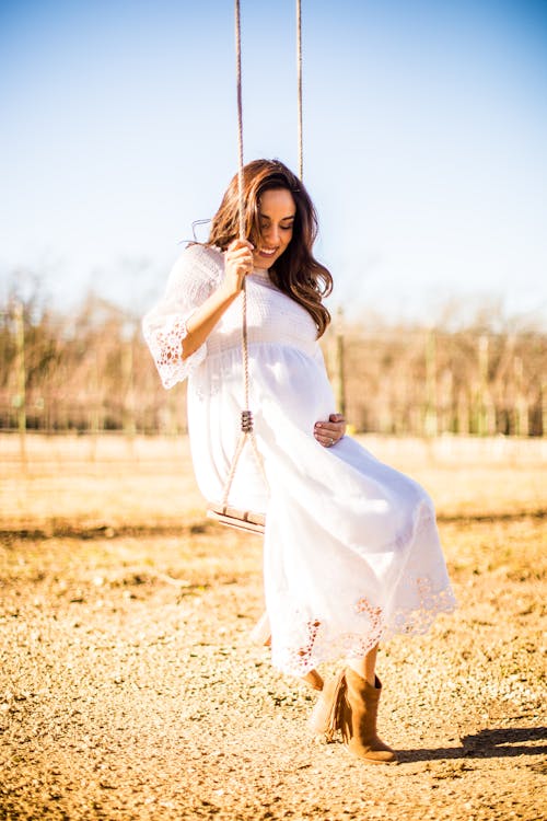 Free A Pregnant Woman in White Dress Looking at Her Baby Bump while Sitting on a Swing Stock Photo