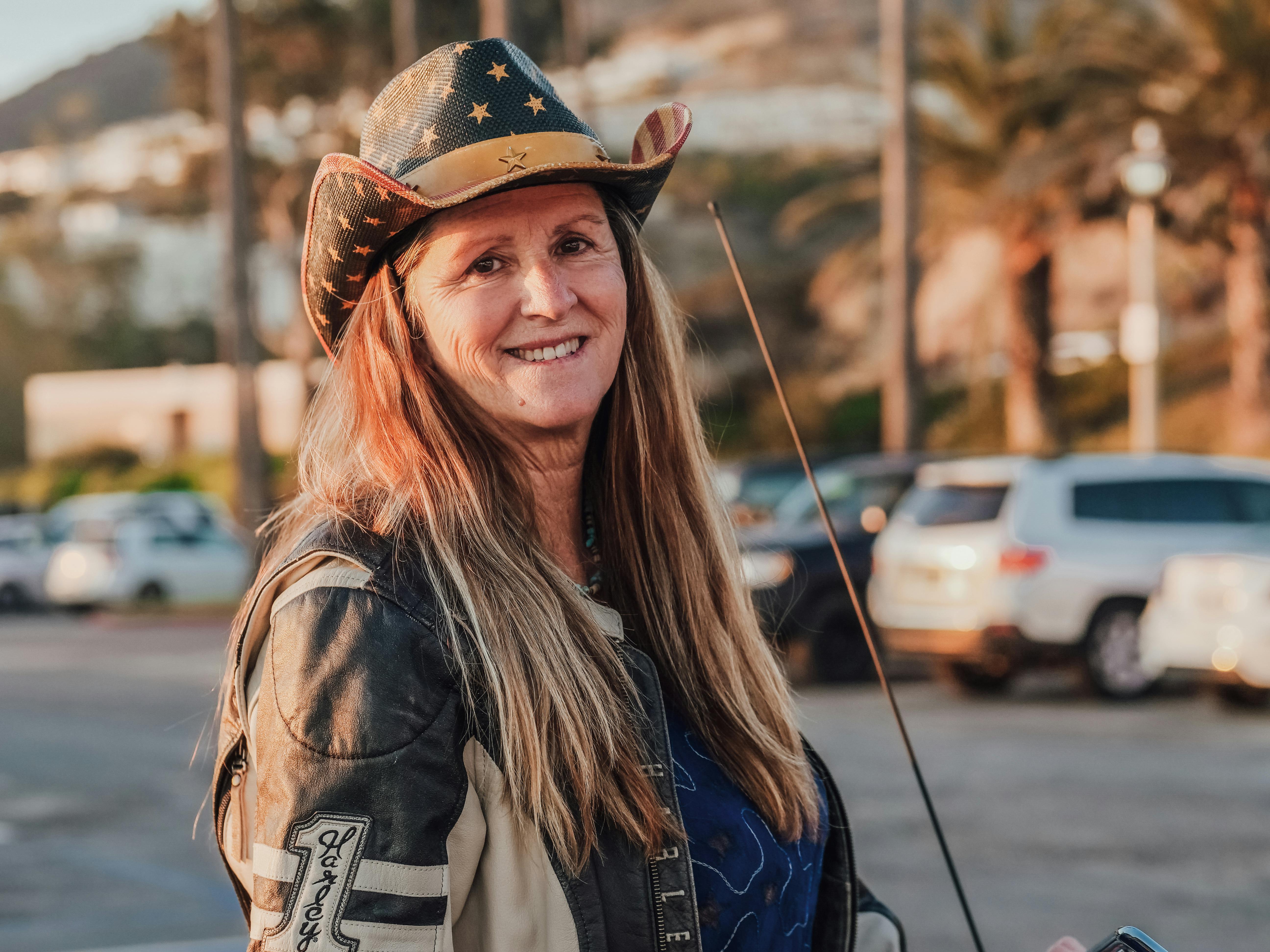 A mature woman wearing a cowboy hat smiles warmly outdoors at sunset.