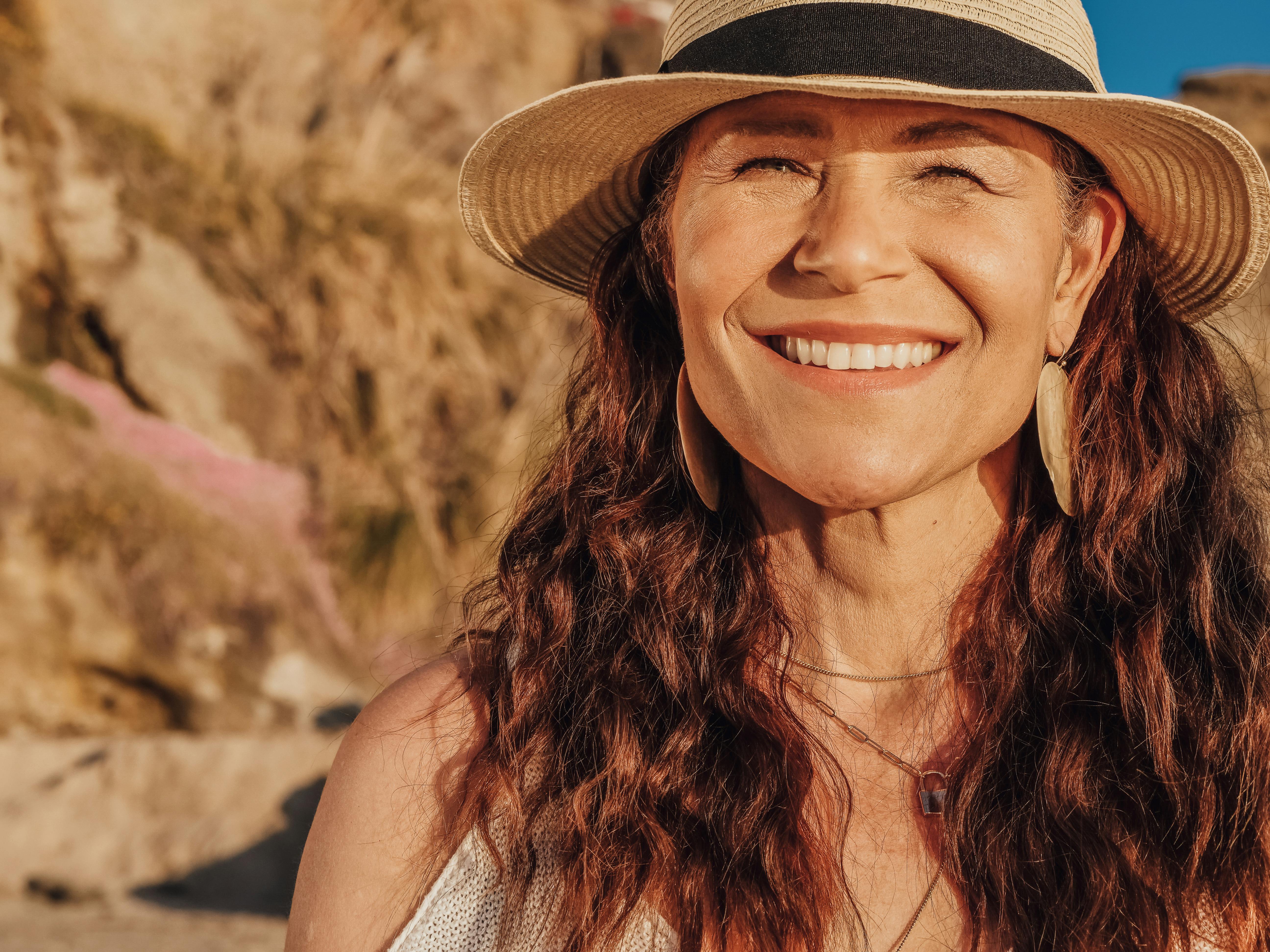 Joyful woman with curly hair and straw hat smiling under the sun on a sandy beach.