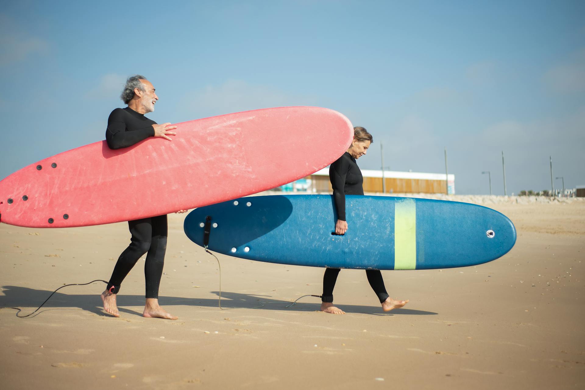 Elderly couple carries surfboards on a sunny Portuguese beach, enjoying an active lifestyle.