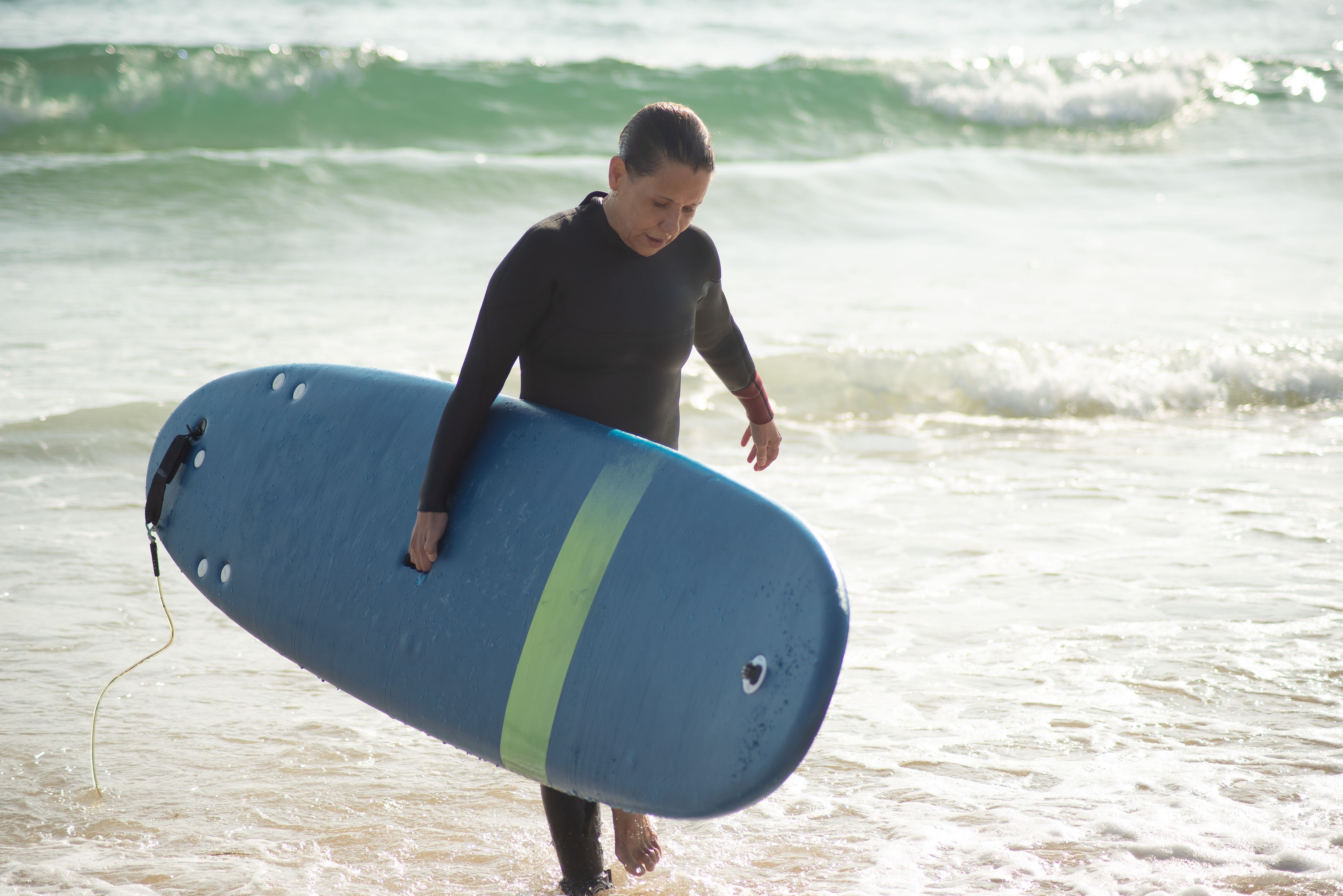 A woman walks on a sunny beach in Portugal, carrying a blue surfboard.