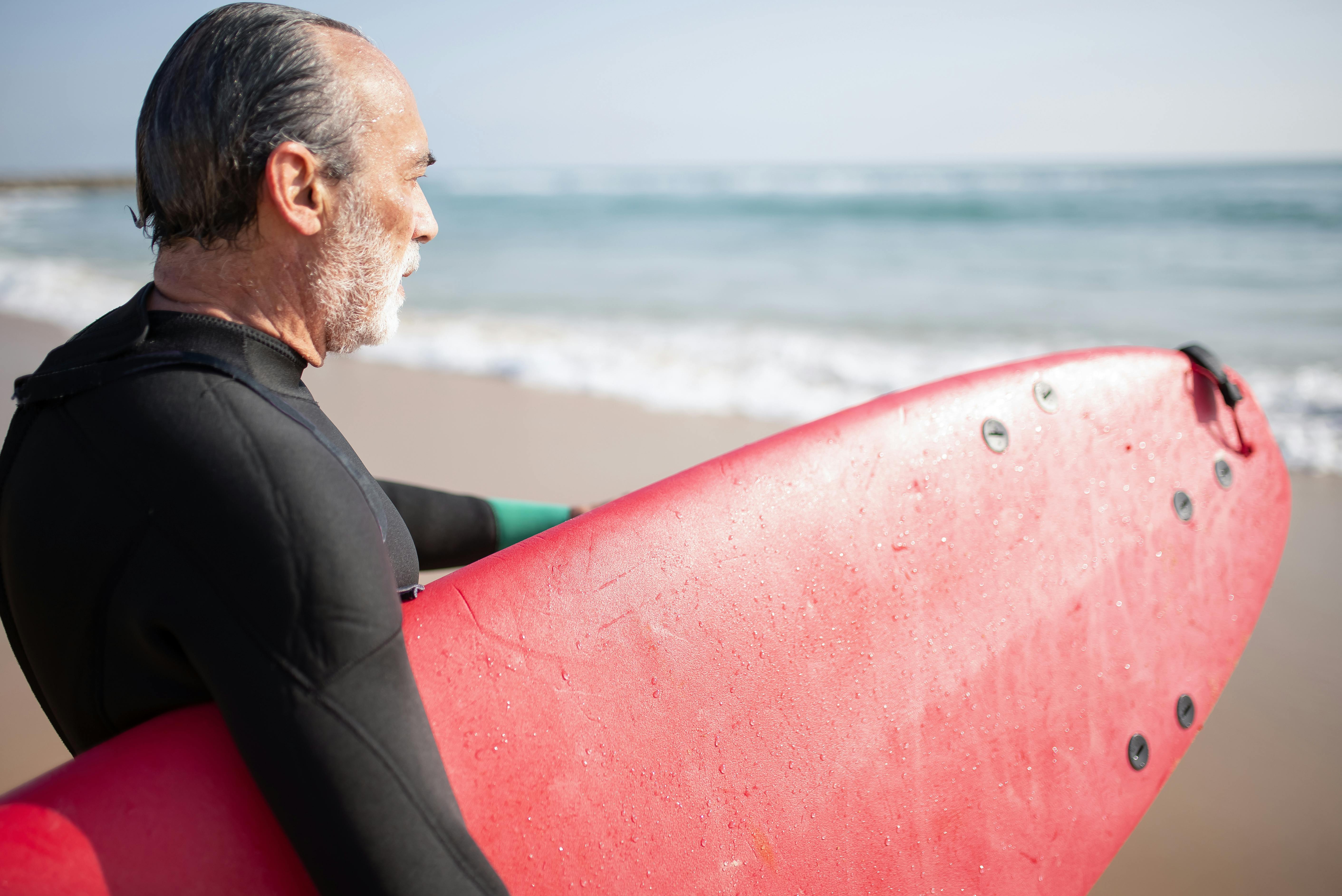 Senior man holding a red surfboard on a sunny beach in Portugal, ready to surf the waves.