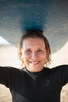 Cheerful elderly woman holding surfboard above head while enjoying sunny day at the beach.