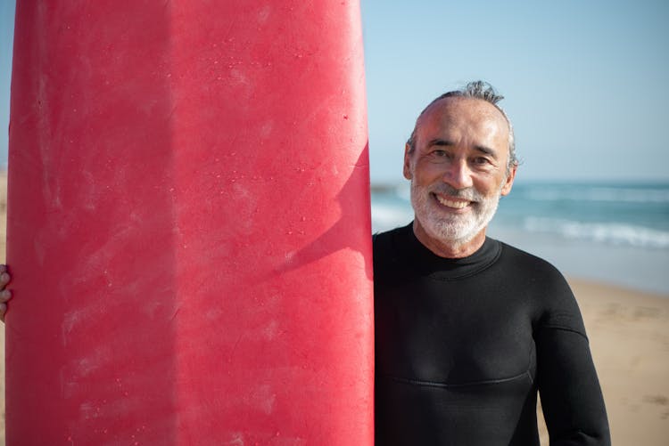 An Elderly Man Wearing Black Rash Guard Smiling At The Camera While Holding Surfboard