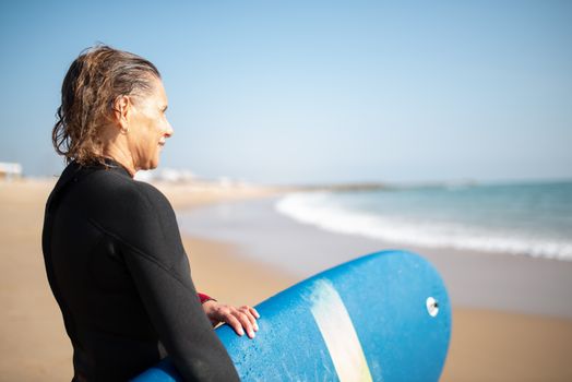 A senior woman holding a blue surfboard while looking at the ocean on a sunny beach in Portugal.