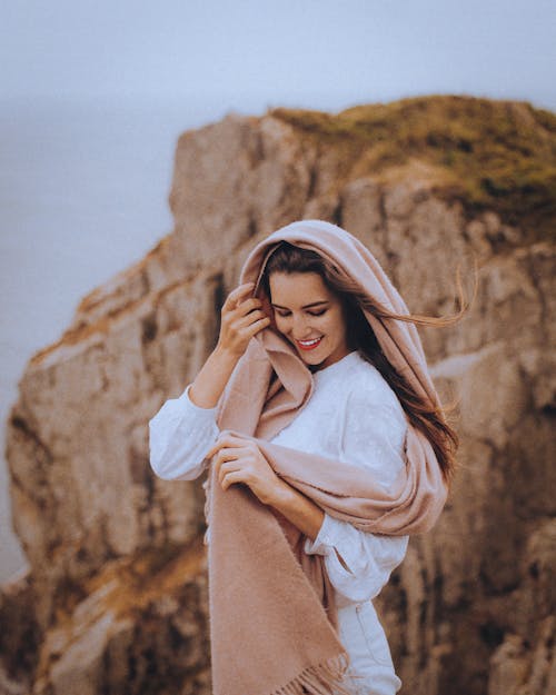 Charming female in casual clothes and scarf standing on rocky cliff in countryside while smiling with closed eyes