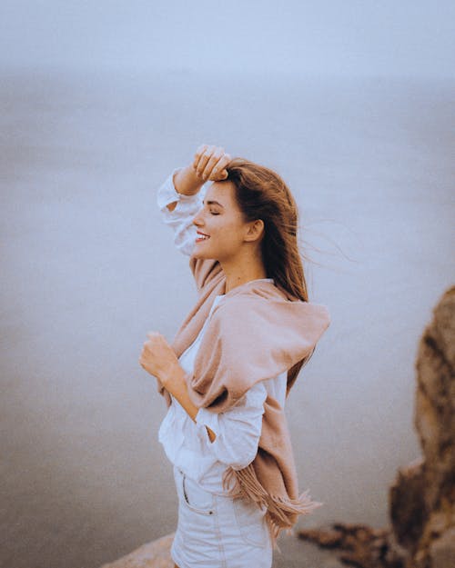 Happy female in stylish outfit standing with closed eyes on cliff above sea while touching face in daylight