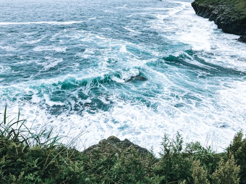 From above of foamy waves of powerful turquoise sea washing rocky formations with green plants in daylight