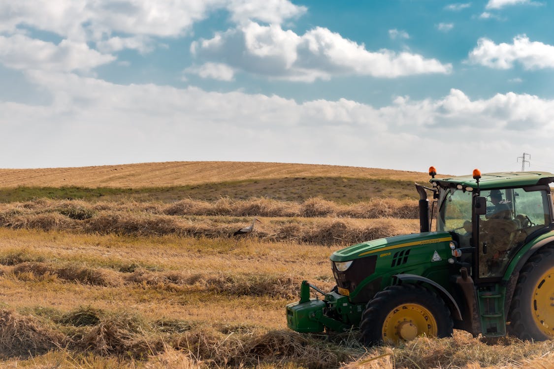 Unrecognizable male farmer driving tractor on dried grassy meadow in countryside near hills under cloudy blue sky in daytime