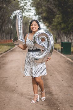 Smiling young woman holding silver number balloons to celebrate her 19th birthday in a park.