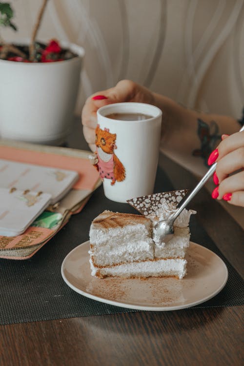 Crop unrecognizable female with delicious cake piece and cup of tea at table in house