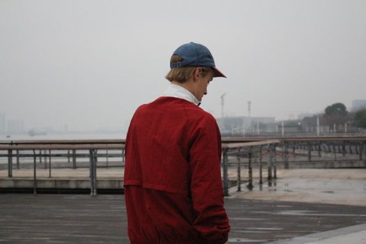 A person in a red jacket and cap looks out over a waterfront on a cloudy day.