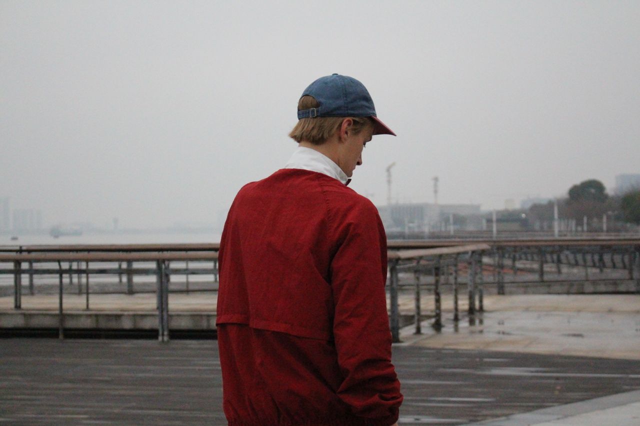 a person in a red jacket and cap looks out over a waterfront on a clou