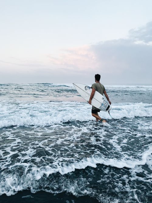 Free A lone surfer carrying a surfboard walks into the waves at Bali beach during a serene day. Stock Photo