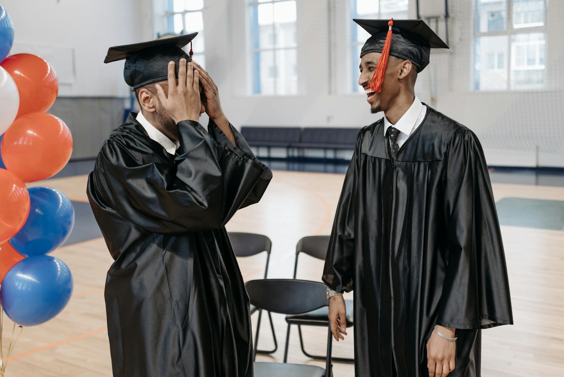Two graduates sharing a joyful moment in caps and gowns indoors.