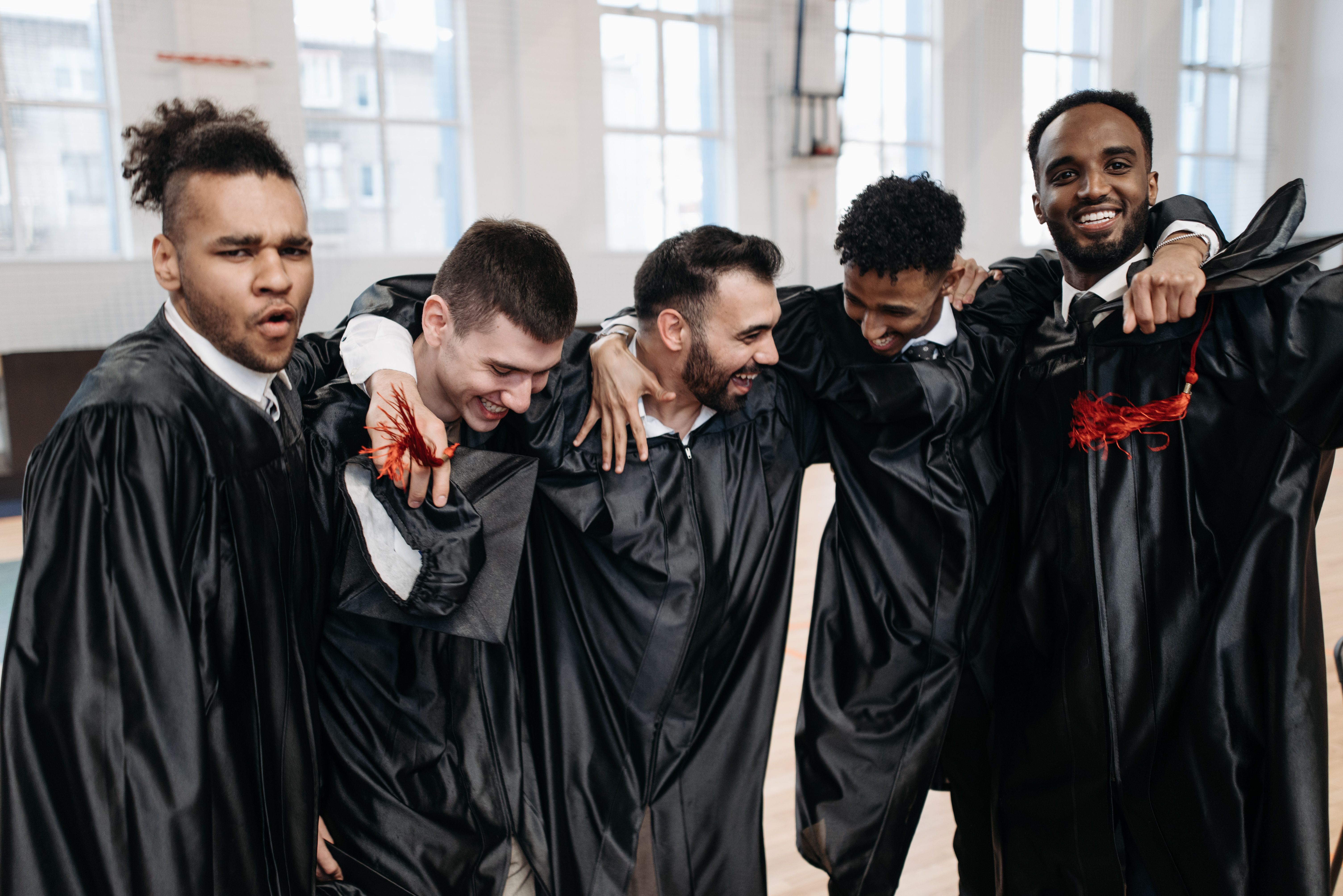 Group of graduates celebrating joyfully in a gymnasium, wearing gowns and smiling.
