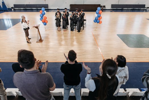 Graduates standing and clapping in a gym after receiving diplomas during a graduation ceremony.
