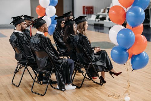 Free A group of graduates in caps and gowns seated indoors with colorful balloons nearby. Stock Photo