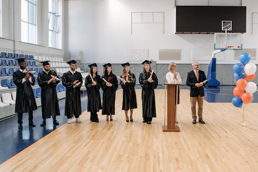 Graduates celebrate in a gymnasium, diverse group in caps and gowns, receiving diplomas.