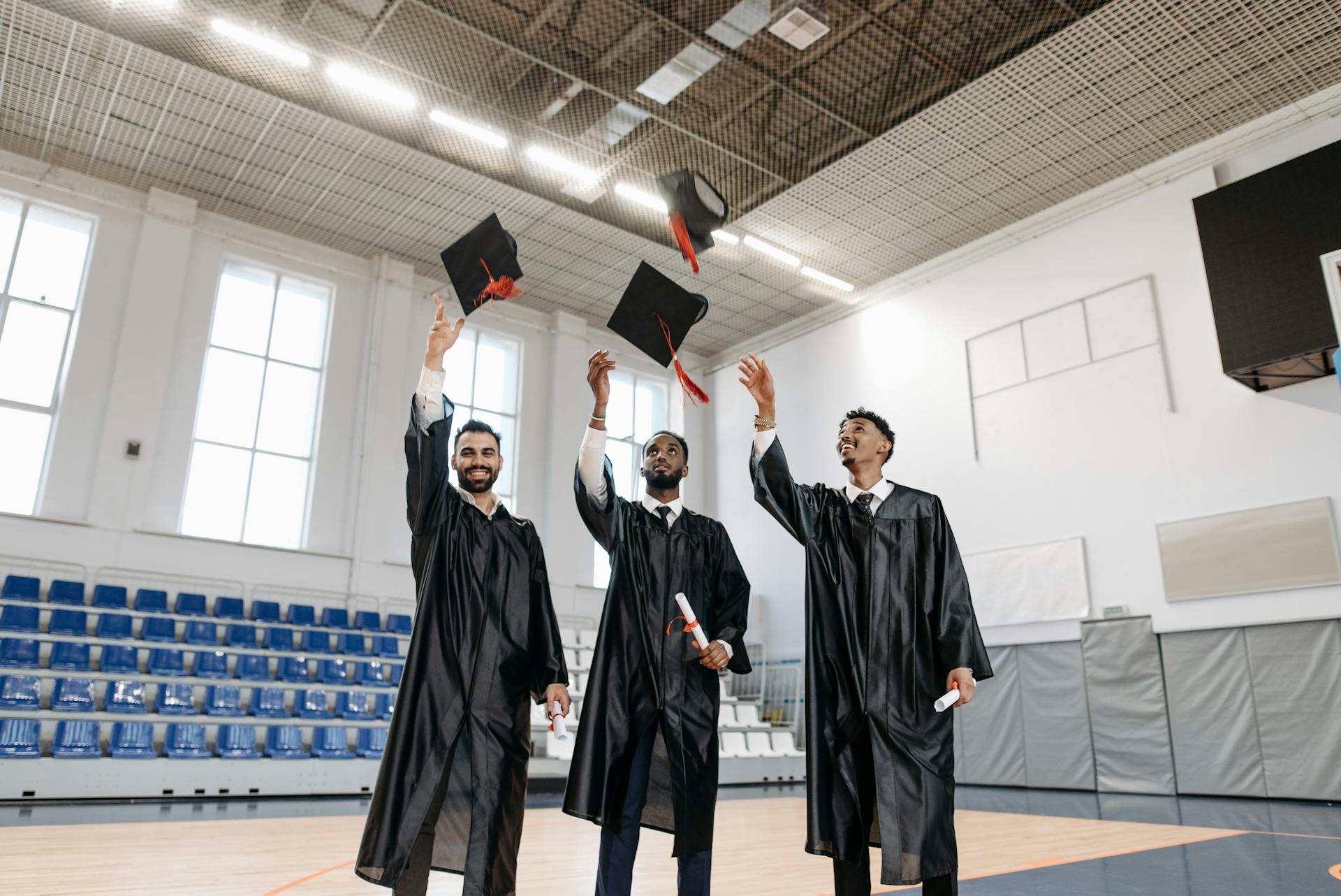 Three graduates celebrate by throwing caps indoors, symbolizing achievement and joy.