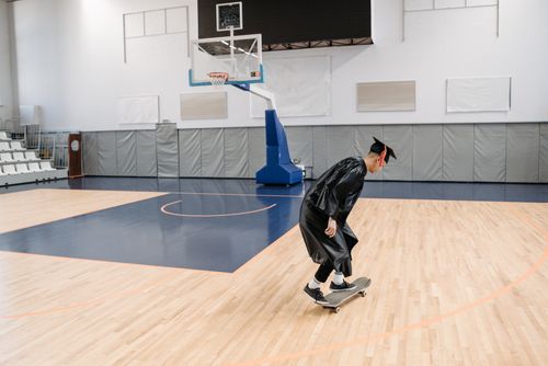 Free A graduate in a cap and gown skateboards on a basketball court indoors. Stock Photo