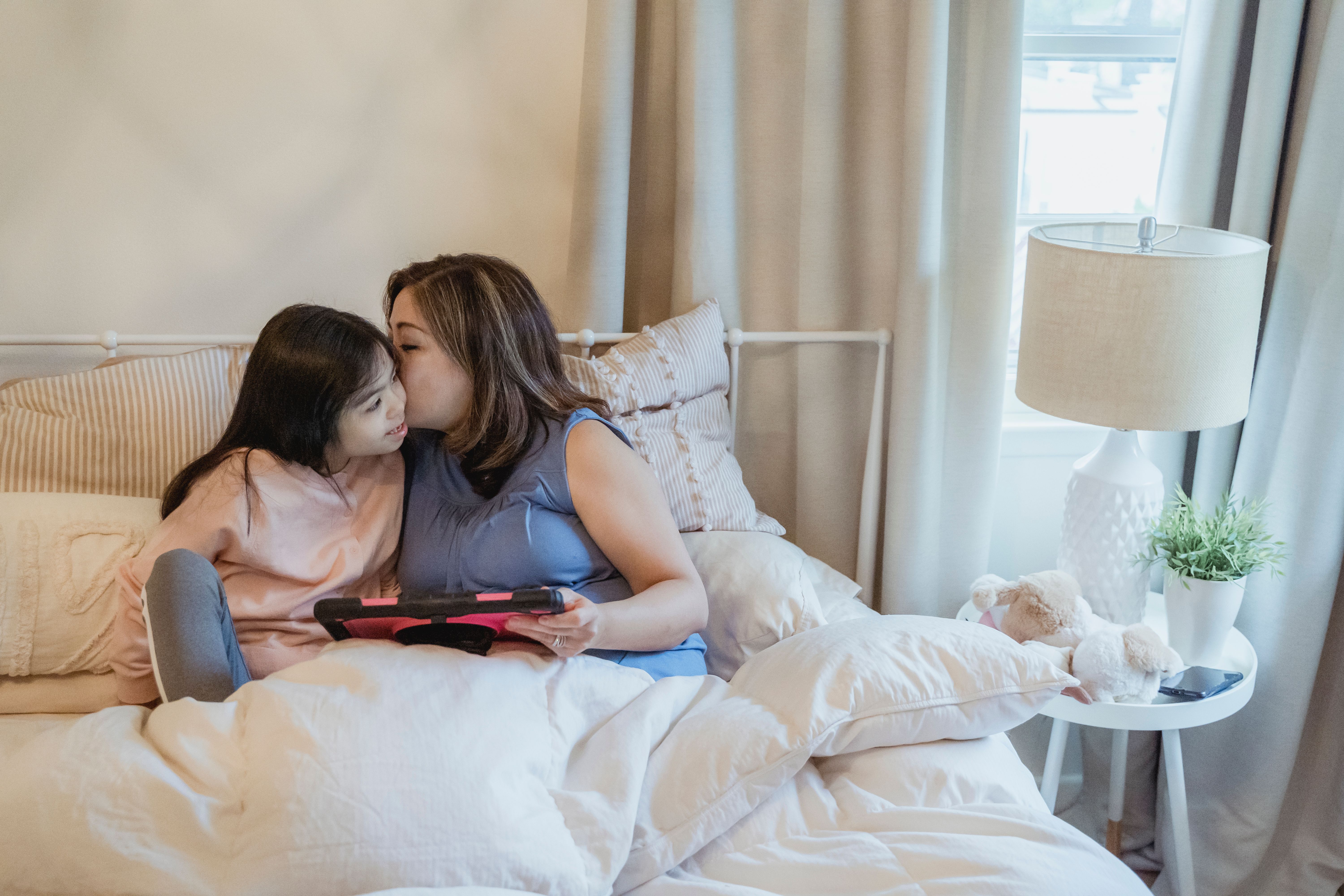 A tender moment of a mother kissing her daughter while sitting on a bed with a tablet.