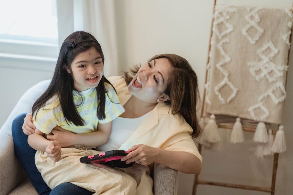 A joyful mother and daughter bonding while using a tablet, seated indoors.