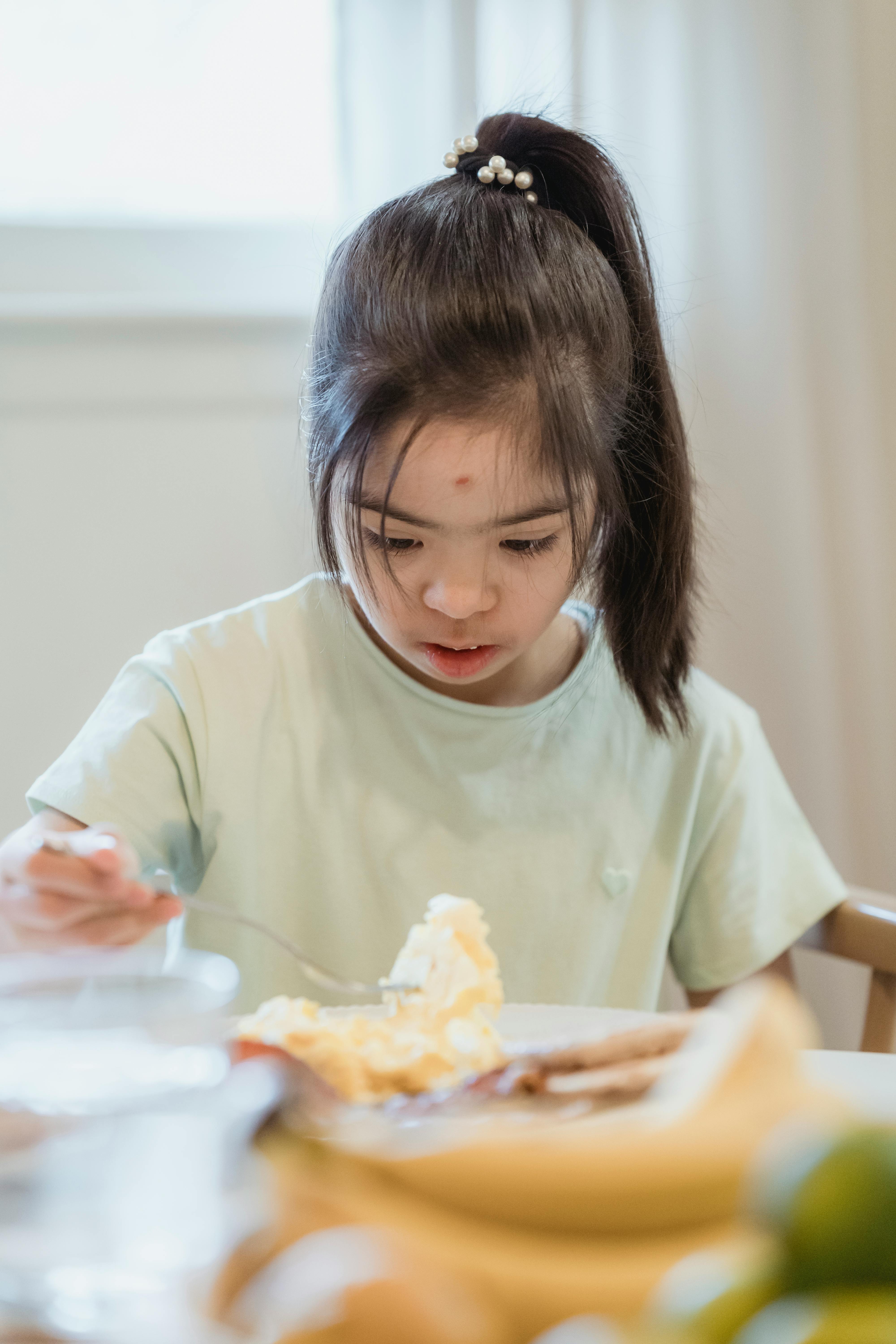 A young girl with Down syndrome enjoys a meal at a table, showcasing innocence and simplicity.