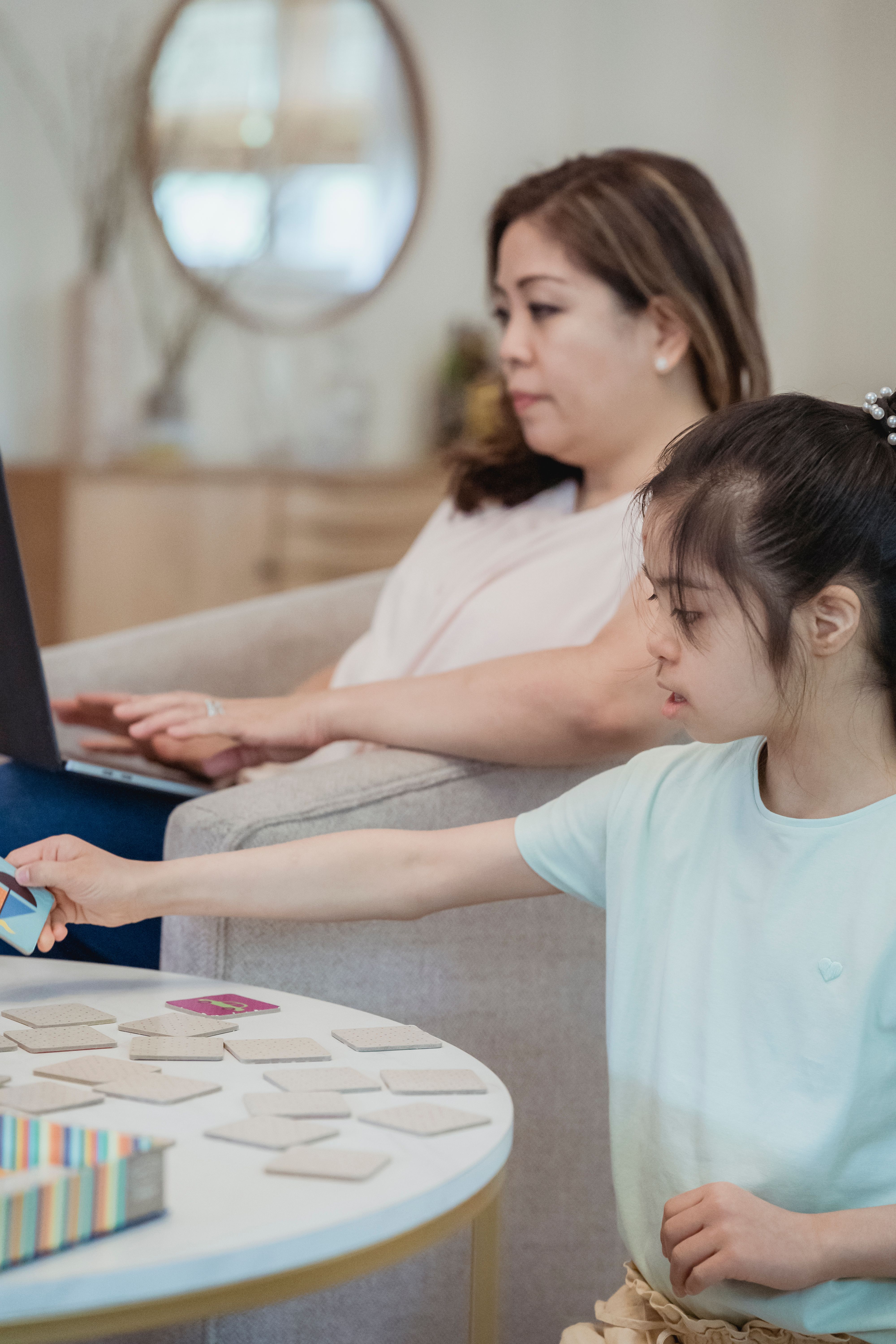 A mother works on her laptop while her daughter plays a memory game in a cozy living room setting.