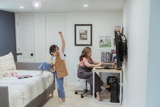 A mother working on a laptop while her daughter plays in a cozy bedroom setting.