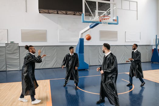 Graduates in black gowns playing basketball indoors, celebrating university success.