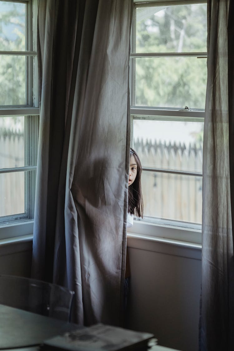 A Girl Hiding Behind A Curtain Near The Window