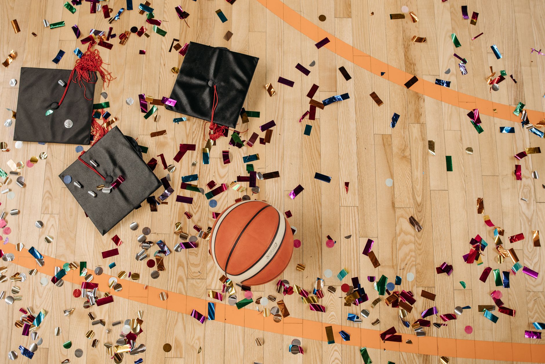 Top view of graduation caps and basketball on confetti-sprinkled gym floor.