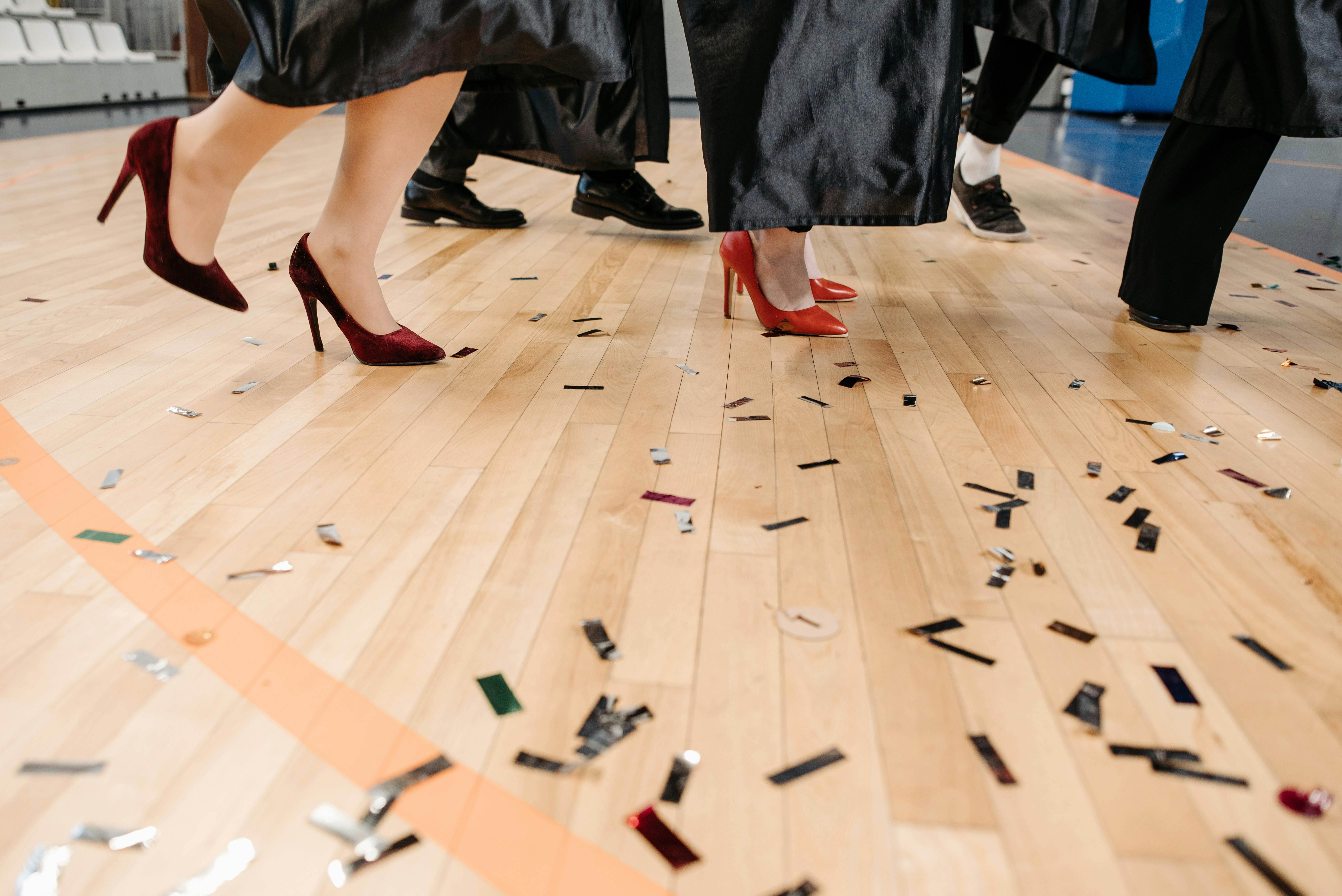 Graduates in gowns and colorful heels walk through scattered confetti on a wooden floor.