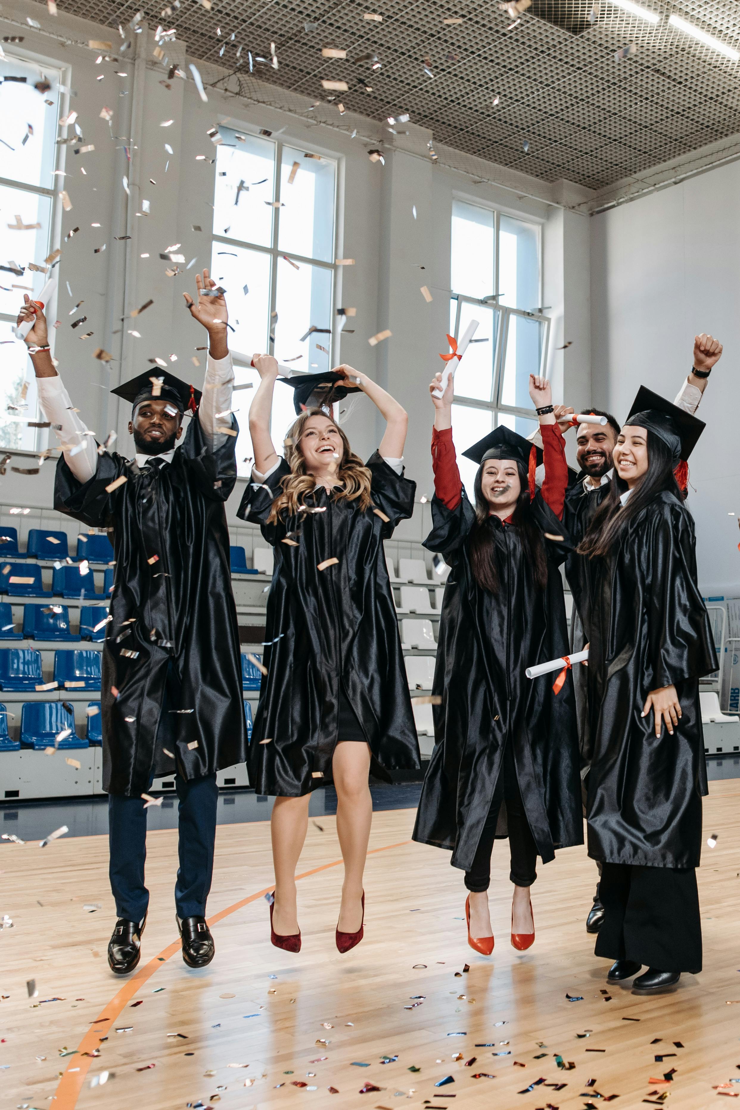 Graduates in gowns rejoice with confetti inside a gymnasium, celebrating success and achievement.