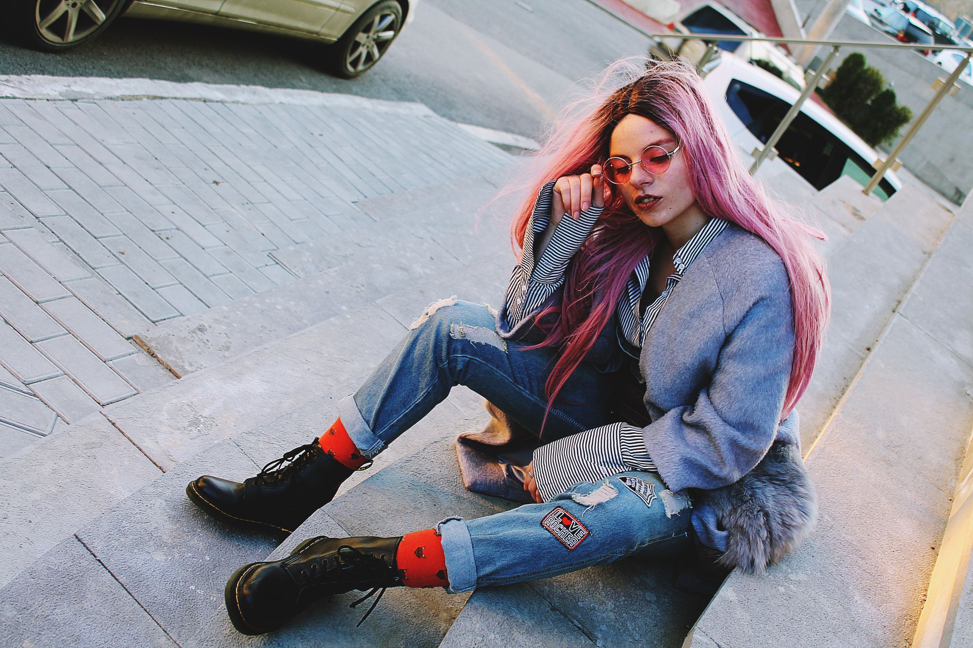 Fashionable woman with pink hair and sunglasses sitting outdoors on steps.