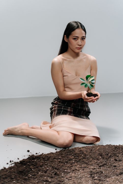 Free A young woman in a beige dress kneeling indoors and holding a small plant, symbolizing environmental conservation. Stock Photo