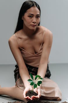 A woman in a spaghetti strap dress holds a small plant in soil, symbolizing eco-consciousness.
