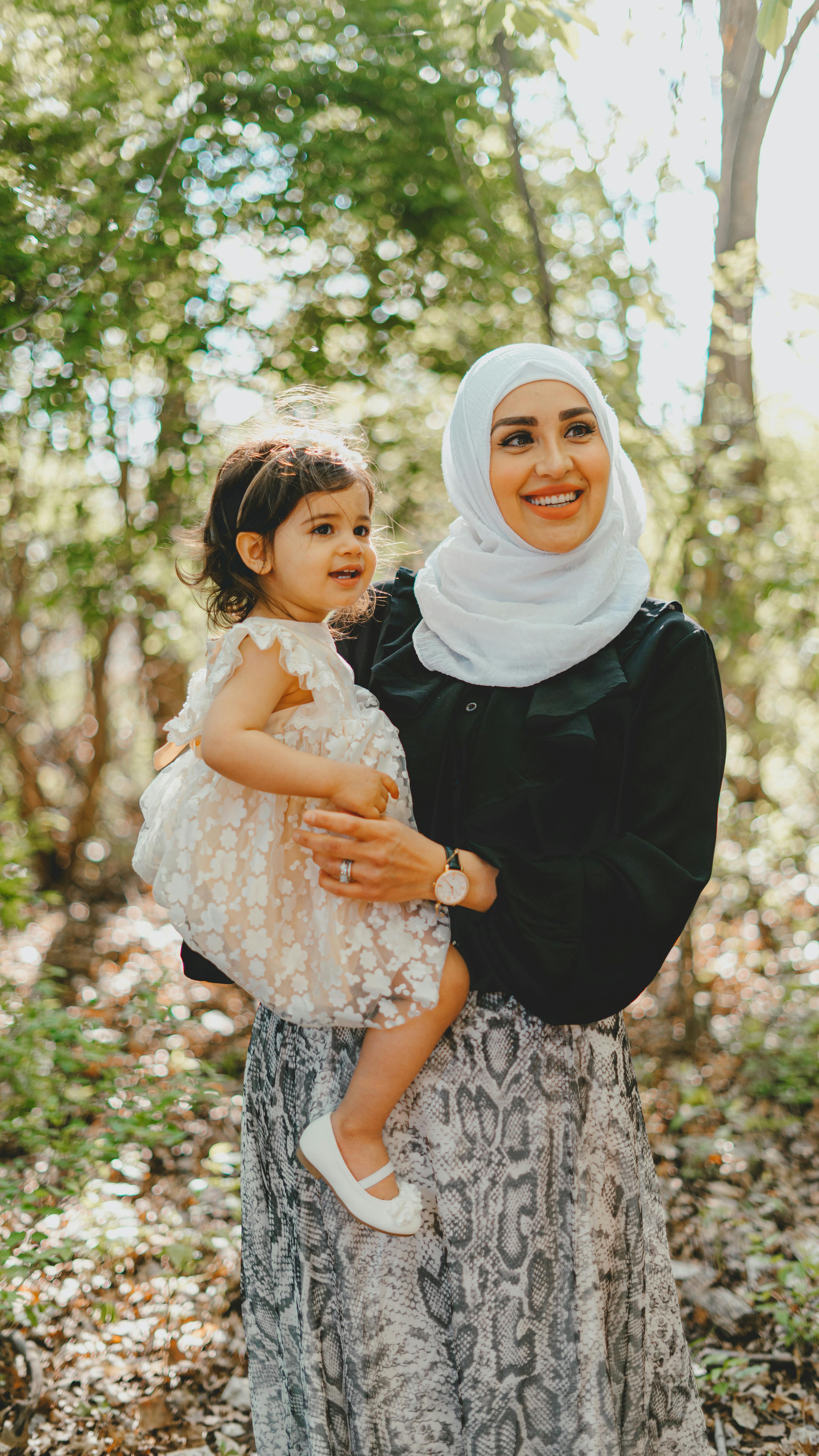 Cheerful Muslim mother in white hijab with cute daughter in arms looking away while standing in park with green trees on summer day